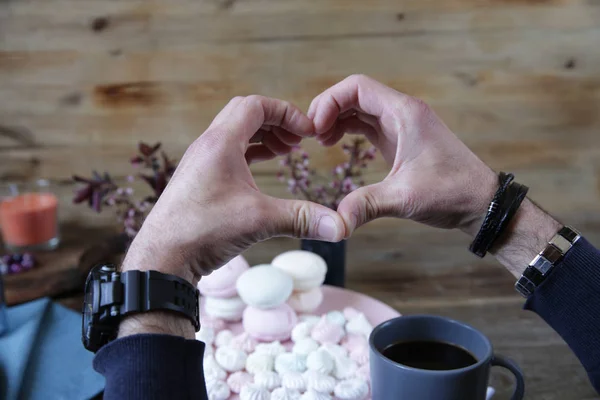 men\'s hands in watches and bracelets folded in the shape of a heart against the background of pink marshmallow, a cup of coffee and spring flowers on a wooden rustic background