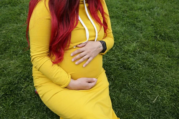 坐在绿色草坪上的一个穿着黄色连衣裙的孕妇的肚子 — 图库照片
