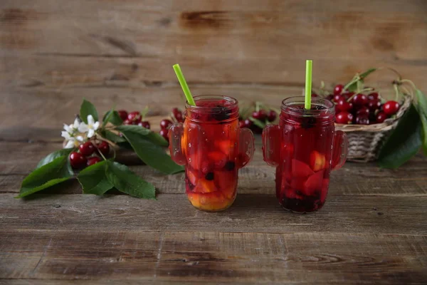 red compote of fresh berries and fruits in a glass jar with a straw for drinking with fresh berries of sweet cherry in a basket on a table made of natural wood. Fresh summer drink. With copy space.