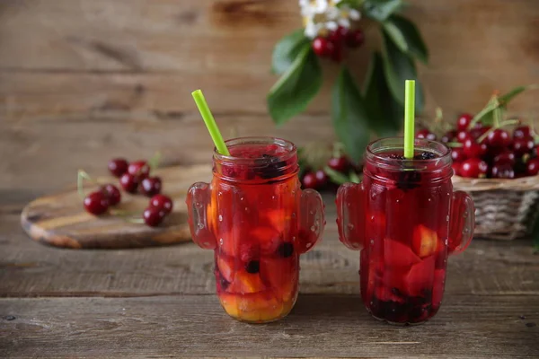 red compote of fresh berries and fruits in a glass jar with a straw for drinking with fresh berries of sweet cherry in a basket on a table made of natural wood. Fresh summer drink. With copy space.