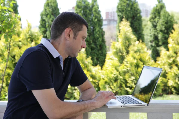 Portrait of a young handsome man with an open laptop in the park on a background of trees with green and yellow foliage with a reflection of trees in his computer. Business and communication.