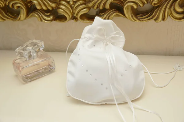 White wedding silk handbag with rhinestones and white ribbon for cosmetics and a bottle of perfume on the dressing table with a gold design. Morning of the bride. Luxury.