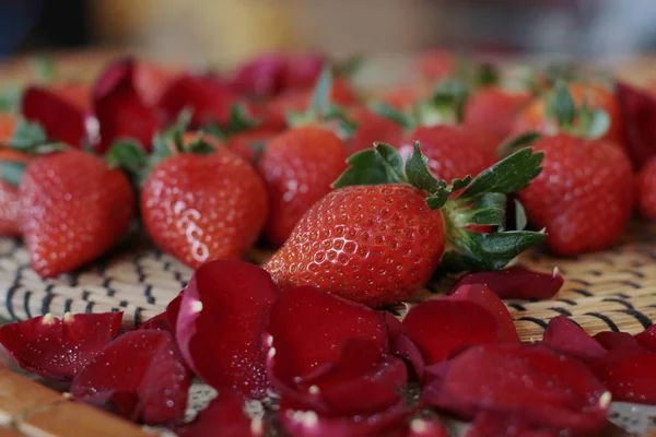 柔らかいぼかしの背景に木製のテーブルの上に赤いバラの花びらと緑の葉と熟した美しい赤いイチゴ — ストック写真