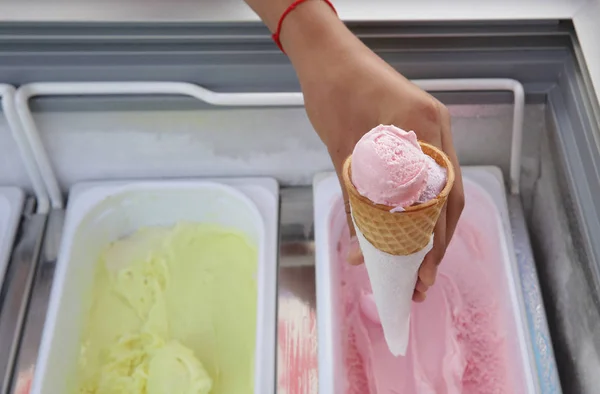 Waffle cone with delicious strawberry ice-cream in the hand. Pink ball of strawberry ice cream in a waffle cone on a background of strawberry and banana ice cream in iron container. Summer.