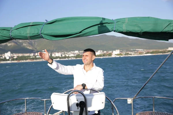 attractive man in white stands at the helm of a yacht at sea and looks at the horizon with his hand raised. Sailing, tourism, travel and people concept. Happy man at the on steering wheel and navigation.