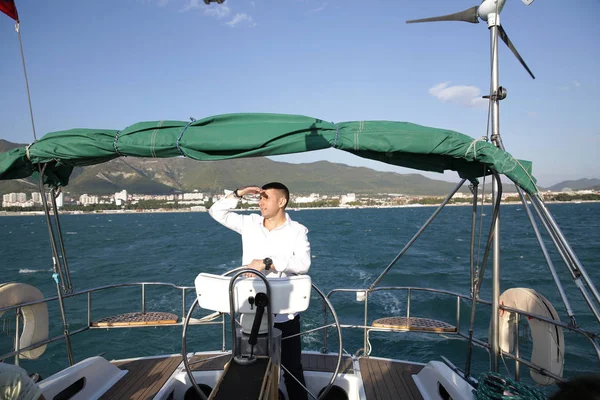 attractive man in white stands at the helm of a yacht at sea and looks at the horizon with his hand raised. Sailing, tourism, travel and people concept. Happy man at the on steering wheel and navigation.