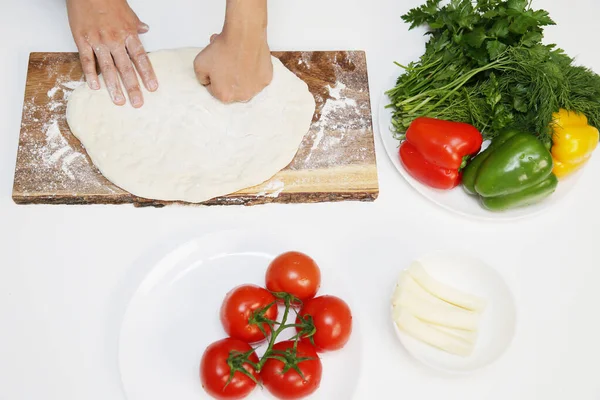 Chef hands prepare pizza dough on a wooden board and ingredients for pizza on the white table. Process of making pizza. Concept of national cuisine.