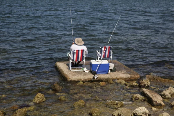 fisherman in a hat sitting on a chair with a red and white stripe is fishing with a fishing rod on the sea or lake there is a container for fish nearby. man sitting with his back.