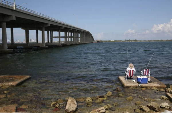 fisherman in a hat sits on a chair with a red and white stripe, fishing with a line, on the sea or on the lake next to a large bridge. man sits with his back.