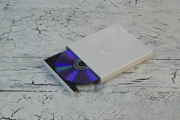 External CD/DVD/Blu-Ray Drive with opened tray and purple Blu-ray (BD) disc on white wooden background (right angle view)