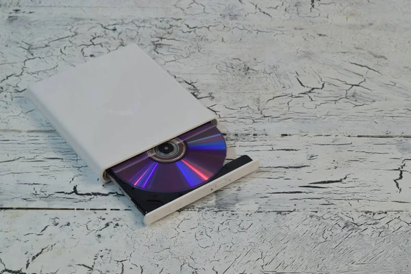 External CD/DVD/Blu-Ray Drive with opened tray and purple Blu-ray (BD) disc on white wooden background (left angle view)
