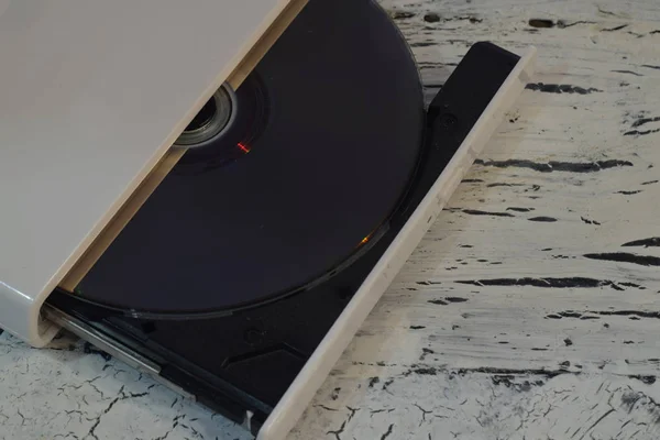 External CD/DVD/Blu-Ray Drive with opened tray and gray Blu-ray (BD) disc on white wooden background (left angle cropped view)