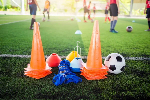 Soccer ball and marker cone with training equipment on green artificial turf with blurry player training background. Soccer Academy.