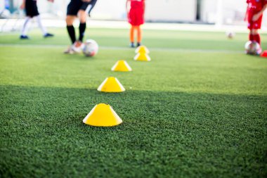 Sarı koni işaretçisine seçici odak yeşil suni sahada futbol eğitim ekipmanları ve bulanık çocuk oyuncuların eğitim geçmişi. Futbol akademisinin eğitim sınıfı için malzeme..