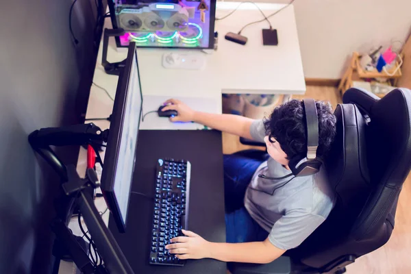 自宅でコンピュータ上でゲームをプレイし ヘッドフォンを着用し 木製のテーブルの上にバックライト付きのカラフルなキーボードを使用してアジアの男の子のゲーマーのトップビューに選択的な焦点 — ストック写真