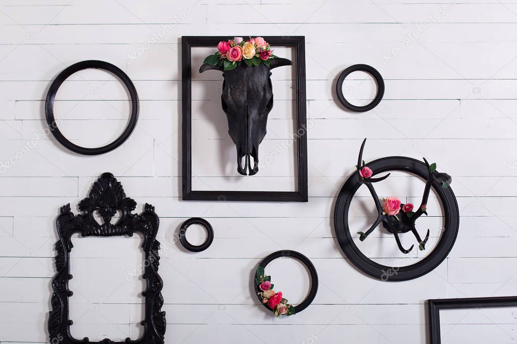 Black skull of a deer and horns on a wooden white wall with empty frames for paintings. The concept of decorating a wooden white wall in the Gothic style in the living room. Decor, vintage, modern