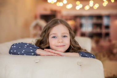 Oturma odasında kanepede oturan gülümseyen küçük kız. Büyük gözleri ve kıvırcık saçları olan ilham verici küçük bir kız. Güzel genç kız kanepede dinleniyor ve kameraya bakıyor. Çocukluk konsepti. Rüya gören çocuk