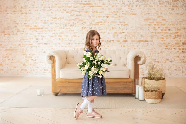 Bir buket yasemin çiçeğiyle gülümseyen kıvırcık saçlı kız rahat bir oturma odasında kanepenin yanında duruyor. İskandinav iç mimarisi. Kutlama. Anneler Günü. Anne için bir buket çiçekli çocuk.. 