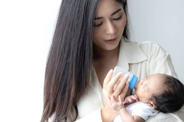Genç Asyalı anne yeni doğmuş şirin bir bebeğe sarılıyor ve beyaz yatak odasında süt şişesini besliyor. Anne ve çocuk kavramı