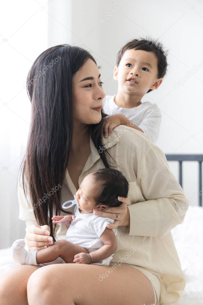Young Asian mother hugging a cute newborn baby sucking fake nipples with the son in white bedroom. Duty of the wife to raise children. Mother and child concept