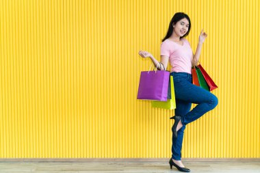 Pembe tişörtlü ve kot pantolonlu Asyalı genç kadın sarı arka planda birkaç kese kağıdı taşıyor. Alışveriş konsepti.