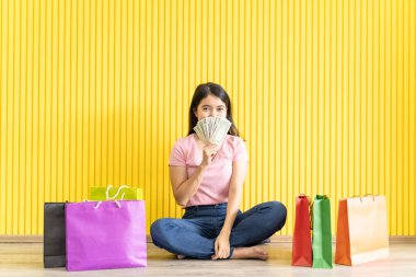 Pembe tişört ve kot pantolon giyen Asyalı genç kadınlar yerde oturuyor ve birkaç kese kağıdıyla dolar banknotları gösteriyor. Sarı arka plan. alışveriş kavramı.