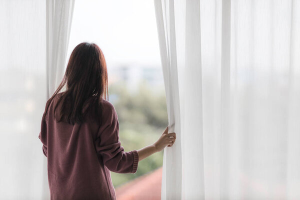 Portrait of beautiful young Asian woman wake up in morning break with freshness and standing near the window and opening white curtains.