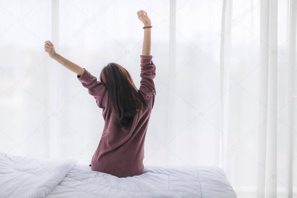 Behind an Asian woman who woke up on a morning break with freshness and she sat on the bed near the window with a white curtain and stretched to keep her body awake.