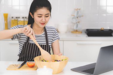 Güzel genç bir kadın internette sağlıklı yemek yapmayı öğreniyor. Beyaz mutfakta bir dizüstü bilgisayardan. Masasında taze sebzeler ve meyveler var..