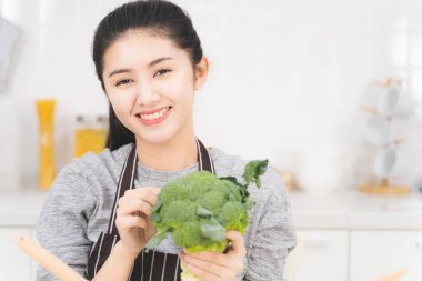 Beautiful asian woman is preparing to cook healthy food which consists of a variety of fruits and vegetables for the family. Portrait of housewife is smiling and holding broccoli. clipart
