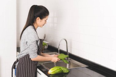 Genç Asyalı ev hanımı sebze yıkıyor mutfakta yemek yapmaya hazırlanıyor..