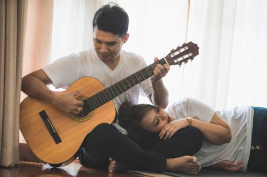Beyaz tişört giyen genç Asyalı çift gitar çalıyor ve sıcak bir evde birlikte şarkı söylüyorlar..