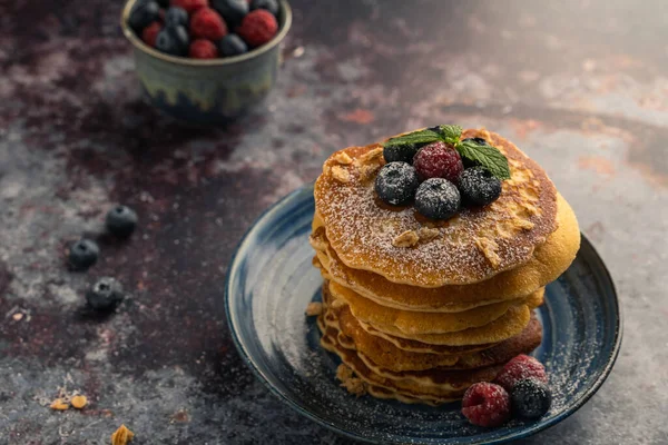 Delicious pancake with blueberries, raspberries and icing sugar in blue dish on rust table.