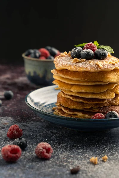 Delicious pancake with blueberries, raspberries and icing sugar in blue dish on rust table.