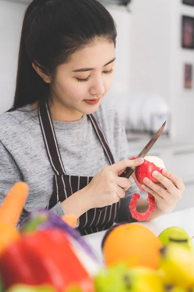 Beautiful asian woman, housewife peeling red apple in white kitchen with plenty of fresh vegetables and fruits on the table.