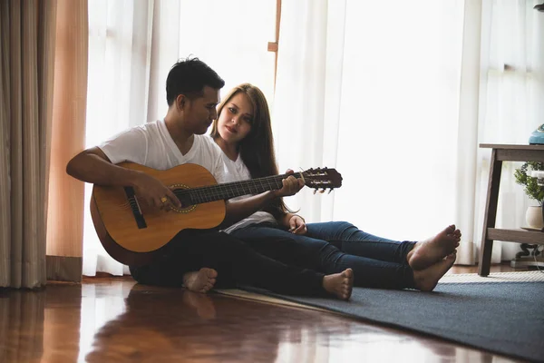 Young Asian couple wearing a white T-shirt is playing the guitar and singing together in a warm home.