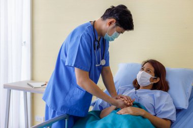 Asyalı erkek doktorlar, hastaları cesaretlendirmek için orta yaşlı kadın hastalarla el ele tutuşup üniforma ve tıbbi maskeler takarlar. Hemşire hastanesi koğuşunda empati ve teşvik.