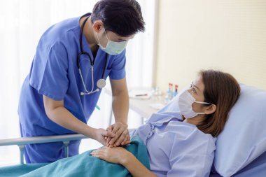 Asyalı erkek doktorlar, hastaları cesaretlendirmek için orta yaşlı kadın hastalarla el ele tutuşup üniforma ve tıbbi maskeler takarlar. Hemşire hastanesi koğuşunda empati ve teşvik.