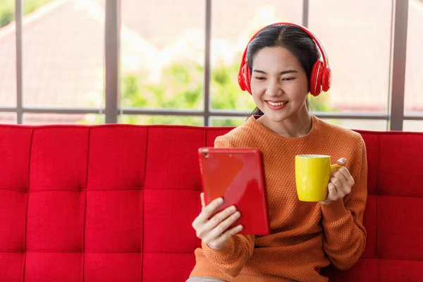 young Asian woman relaxing on a red sofa inside the house. Women drinking coffee and listening music from tablet. work from home.