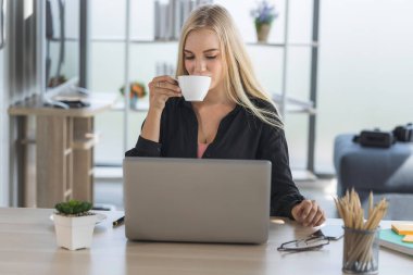 Sabah güneşiyle ev ofisinden çalışan genç bir iş kadını. Kadın kahve içiyor ve sosyal ağda iletişim kurmak ve internetten bilgi aramak için laptop kullanıyor..