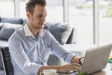 Ev ofisinden sabah güneşiyle çalışan genç bir adamın portresi. Sosyal ağda iletişim kuran ve çevrimiçi bilgi arayan mutlu adam.