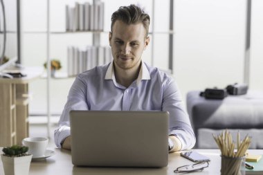 Ev ofisinden sabah güneşiyle çalışan genç bir adamın portresi. Sosyal ağda iletişim kuran ve çevrimiçi bilgi arayan mutlu adam.