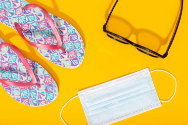 Düz kompozisyon, yaz tatili aksesuarları ve sarı arka planda yüz maskesi. Coronavirus covid-19 salgını sırasında yaz ya da tatil konsepti. Üst görünüm