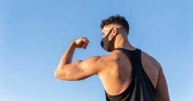 Mavi gökyüzü arka planında kaslarını gösteren yüz maskesi takan atletik beyaz bir adam. COVID-19 Coronavirus 'a karşı savaş kavramı. Boşluğu kopyala