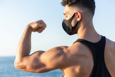 Mavi gökyüzü arka planında kaslarını gösteren yüz maskesi takan atletik beyaz bir adam. COVID-19 Coronavirus ile mücadele kavramı