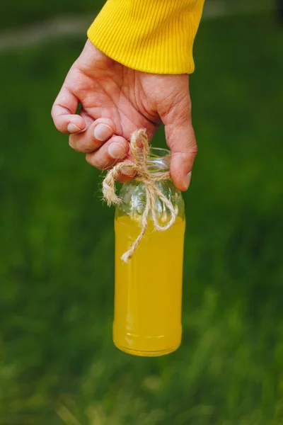 Man holds in his hand bottle of homemade lemonade on the background of trees in the park and green grass. sunny day, summer. closeup. picnic healthy food fresh. cropped image — Stock Photo, Image