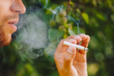 için için yanan sigara doğa yeşil ağaçlar arka planı üzerinde bir sakallı bir adam elinde. Nikotin tütün dumanı. Sağlıksız yaşam tarzı. Yakın çekim, ash. İçme molası