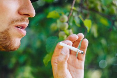 için için yanan sigara doğa yeşil ağaçlar arka planı üzerinde bir sakallı bir adam elinde. Nikotin tütün dumanı. Sağlıksız yaşam tarzı. Yakın çekim, ash. İçme molası