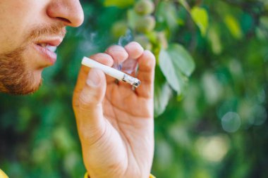 için için yanan sigara doğa yeşil ağaçlar arka planı üzerinde bir sakallı bir adam elinde. Nikotin tütün dumanı. Sağlıksız yaşam tarzı. Yakın çekim, ash. İçme molası