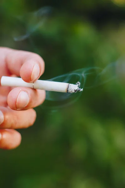 smoldering cigarette in a man\'s hand in nature against the backdrop of green trees. Nicotine tobacco smoke. Unhealthy Lifestyle. Close up ash. Harmful habit for health. Environmental pollution.